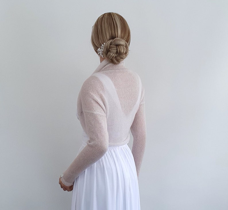 May include: A sheer, light pink knitted shrug with long sleeves. The shrug is draped over the shoulders and arms of a person wearing a white dress.