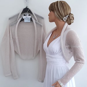 May include: A light beige knitted shrug with long sleeves. The shrug is draped over a hanger and a mannequin is wearing a similar shrug.