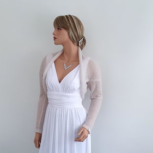 May include: A light beige knitted shrug with long sleeves. The shrug is draped over the shoulders of a mannequin wearing a white dress.