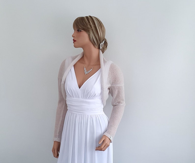 May include: A light beige knitted shrug with long sleeves. The shrug is draped over the shoulders of a mannequin wearing a white dress.