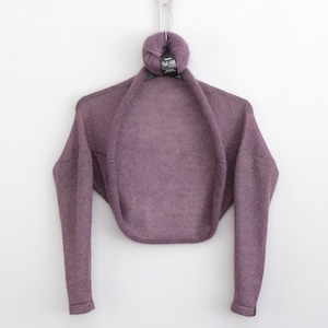 May include: A purple knitted shrug with long sleeves. The shrug is hanging on a hanger with a ball of yarn on top.