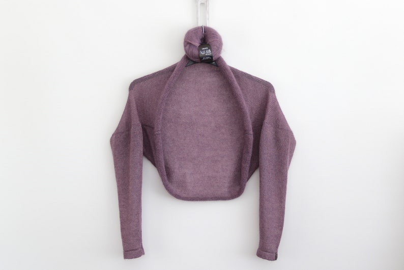 May include: A purple knitted shrug with long sleeves. The shrug is hanging on a hanger with a ball of yarn on top.