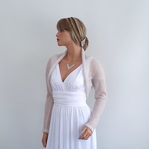 May include: A light beige knitted shrug with long sleeves. The shrug is draped over the shoulders of a mannequin wearing a white dress.