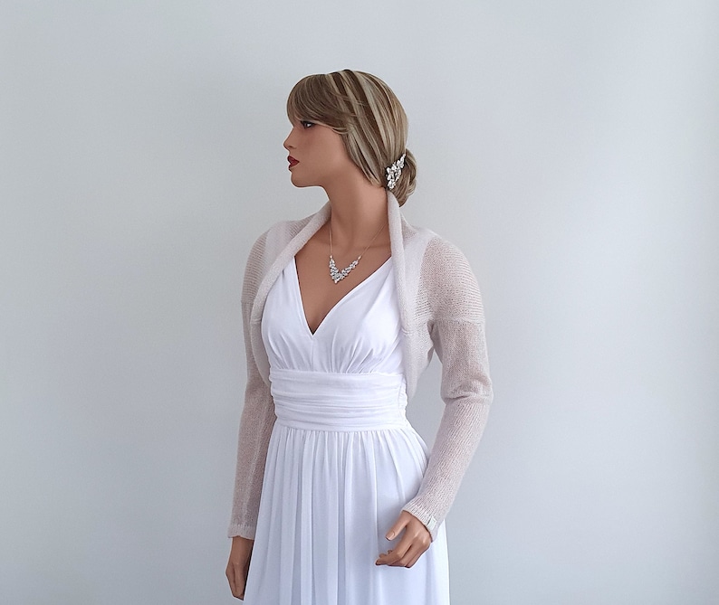 May include: A light beige knitted shrug with long sleeves. The shrug is draped over the shoulders of a mannequin wearing a white dress.