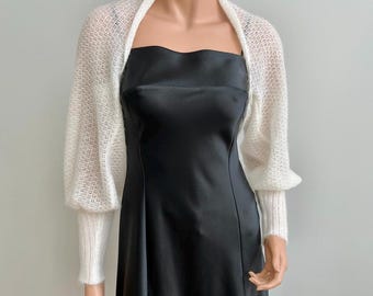 May include: A white knitted shrug with long sleeves and a delicate openwork pattern. The shrug is worn over a black satin dress.