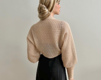 May include: A beige knitted shrug with long sleeves and a loose fit. The shrug is made of a lightweight, airy fabric and has a delicate, open-weave pattern.