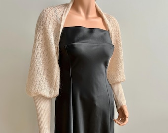 May include: A cream-colored knitted shrug with long sleeves and a loose fit. The shrug is made of a soft, lightweight yarn and has a delicate, openwork pattern.
