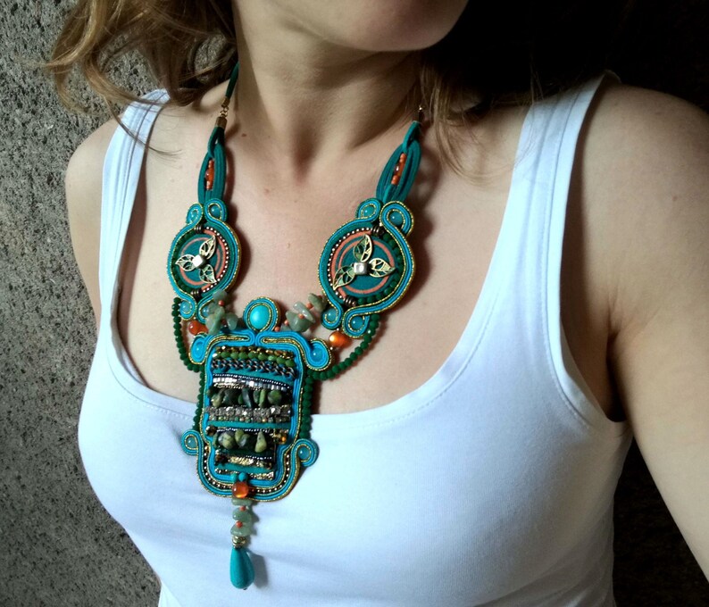 May include: A turquoise and green statement necklace with a large, intricate pendant featuring a geometric design. The pendant is adorned with turquoise and green stones, gold accents, and beads.
