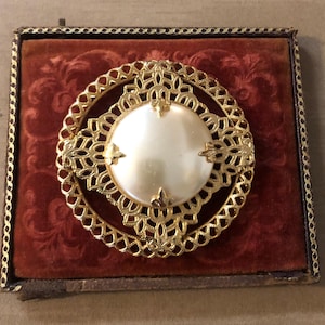 May include: A circular gold-tone brooch featuring an intricate filigree metal frame surrounding a central smooth white faux pearl cabochon. The brooch is displayed on a square, dark red velvet-lined box with a decorative gold-tone border trim.