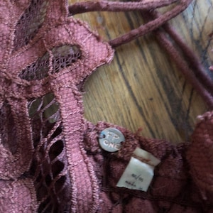 May include: Close-up of a burgundy-colored lace top with a crisscross back. The top features a floral lace design and a small metal tag with the letters "FP ONE". The tag is attached to the top.