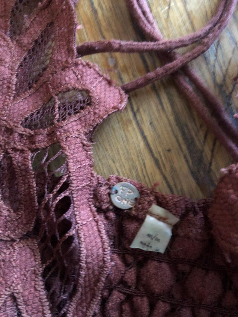 May include: Close-up of a burgundy-colored lace top with a crisscross back. The top features a floral lace design and a small metal tag with the letters "FP ONE". The tag is attached to the top.