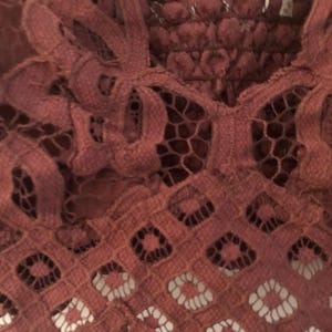 May include: Close-up of a burgundy lace top with intricate floral and geometric patterns. The top features a scalloped edge and delicate straps, showcasing a detailed, openwork design. The fabric appears soft and textured.