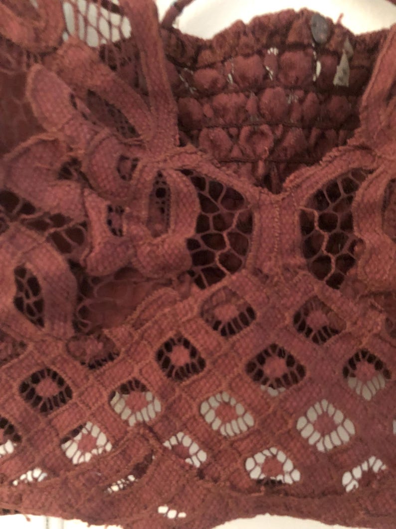 May include: Close-up of a burgundy lace top with intricate floral and geometric patterns. The top features a scalloped edge and delicate straps, showcasing a detailed, openwork design. The fabric appears soft and textured.