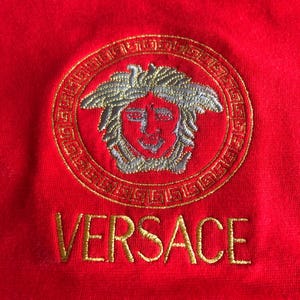 Peut inclure: Gros plan sur un v&ecirc;tement rouge avec un motif circulaire brod&eacute; en or repr&eacute;sentant une t&ecirc;te de M&eacute;duse. Le mot "VERSACE" est brod&eacute; en or en dessous.
