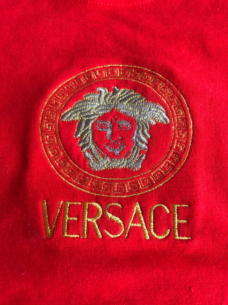 Peut inclure: Gros plan sur un v&ecirc;tement rouge avec un motif circulaire brod&eacute; en or repr&eacute;sentant une t&ecirc;te de M&eacute;duse. Le mot "VERSACE" est brod&eacute; en or en dessous.