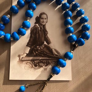 May include: A strand of bright blue beads with silver-colored spacers, draped over a vintage black and white photograph of a woman. The beads have a glossy finish and are strung on a dark cord, with decorative gold-toned chain and coin charms.