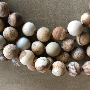 May include: Close-up of a necklace featuring three strands of large, round beads. The beads are primarily beige and brown with unique patterns, strung together with copper-colored wire. The necklace is displayed against a neutral background.