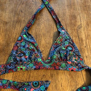 May include: A halter-neck bikini top featuring a dark background with a vibrant, multi-coloured floral and paisley pattern in shades of neon green, bright blue, and coral red. The garment includes triangular cups and long ties for closure.
