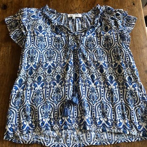 May include: A short-sleeved top with a blue and white paisley pattern. The top has a ruffled neckline and sleeves, a V-neck with a tassel tie, and a slightly flared hem. The fabric appears to be lightweight.