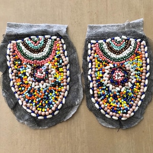 May include: A pair of oval-shaped fabric patches featuring intricate beadwork in a vibrant palette of pink, blue, yellow, green, and white. The beads are arranged in concentric patterns on a black mesh base with a grey fabric top border.