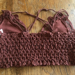 May include: A maroon-colored crop top with a textured, smocked bodice and delicate lace detailing. The top features adjustable spaghetti straps and a soft, layered design. The garment is made of a soft, breathable fabric.