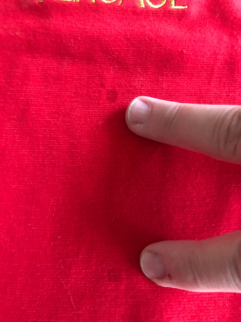 Peut inclure: Gros plan sur un tissu rouge avec le mot "VERSACE" brod&eacute; en fil d'or en haut. Le tissu a un aspect l&eacute;g&egrave;rement textur&eacute;. Un doigt touche le tissu.