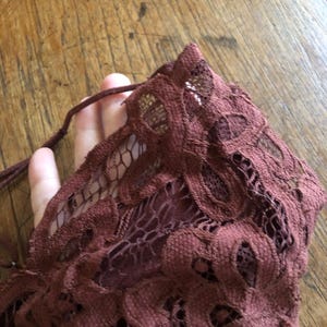 May include: Close-up of a burgundy lace fabric with a floral pattern. The delicate material is gathered, revealing the intricate openwork design. A thin, matching cord is visible, suggesting a tie or drawstring detail.