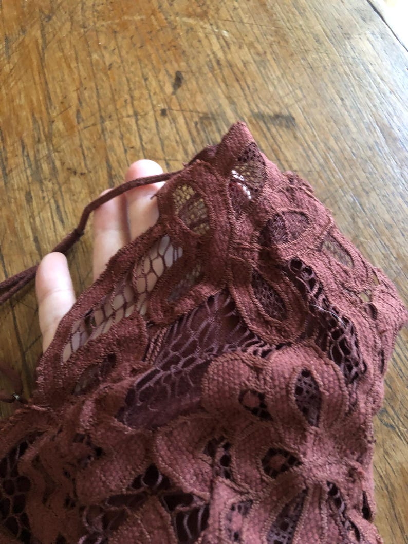 May include: Close-up of a burgundy lace fabric with a floral pattern. The delicate material is gathered, revealing the intricate openwork design. A thin, matching cord is visible, suggesting a tie or drawstring detail.