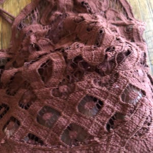 May include: Close-up of a maroon-colored lace top. The intricate lace design features a repeating pattern of openwork shapes. The fabric appears soft and delicate, with a slightly textured surface. The top has thin straps.