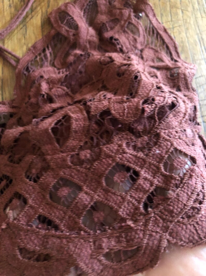 May include: Close-up of a maroon-colored lace top. The intricate lace design features a repeating pattern of openwork shapes. The fabric appears soft and delicate, with a slightly textured surface. The top has thin straps.