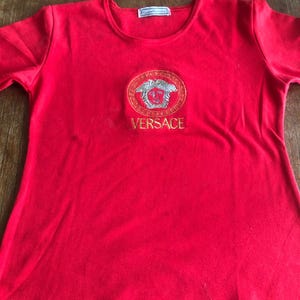 Peut inclure: Un t-shirt rouge vif avec un logo Versace brod&eacute; en or et argent. Le logo ovale est centr&eacute;, avec le nom de la marque en lettres dor&eacute;es. Le t-shirt a des manches courtes et un col rond. Le tissu semble doux.