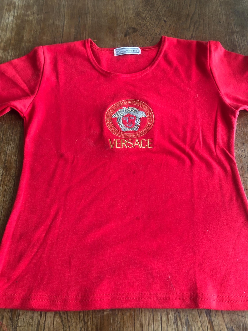 Peut inclure: Un t-shirt rouge vif avec un logo Versace brod&eacute; en or et argent. Le logo ovale est centr&eacute;, avec le nom de la marque en lettres dor&eacute;es. Le t-shirt a des manches courtes et un col rond. Le tissu semble doux.