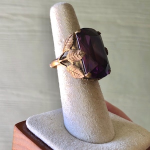 May include: A ring featuring a large, rectangular, deep purple faceted gemstone set in a metallic band. The setting is adorned with detailed, textured metal leaves that extend from the sides of the stone, wrapping around the band's shank.