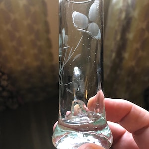 May include: A clear glass bud vase with a cylindrical body and a flared base. The glass features a delicate etched floral design on the front surface, showcasing thin stems and petal details. The vessel is transparent and smooth in texture.