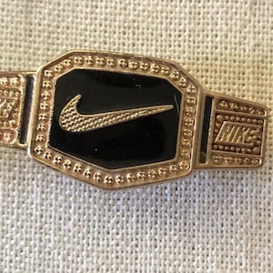 Może przedstawiać: Złota metalowa przypinka w kształcie pasa mistrzowskiego, z centralnym czarnym prostokątnym panelem i teksturowanym złotym logo Nike swoosh. Boczne płytki są wytłoczone napisem NIKE, a kropkowany wzór otacza środkową część przypinki.