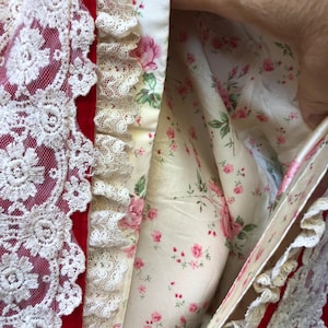 Op de afbeelding: Een rode fluwelen jurk met een witte kantversiering en een bloemenvoering. De voering is crèmekleurig met roze rozen en groene bladeren.