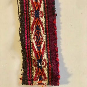 Peut inclure: Textile rectangulaire tissé avec un motif de losange central rouge, orange, blanc et bleu. Les bords sont finis avec une frange marron foncé et magenta. Le textile a un aspect artisanal traditionnel.