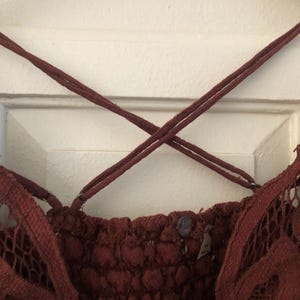 May include: Close-up of a burgundy-colored lace top with crisscross spaghetti straps. The top features a textured, patterned bodice and delicate lace detailing along the edges. A small button and tag are visible.