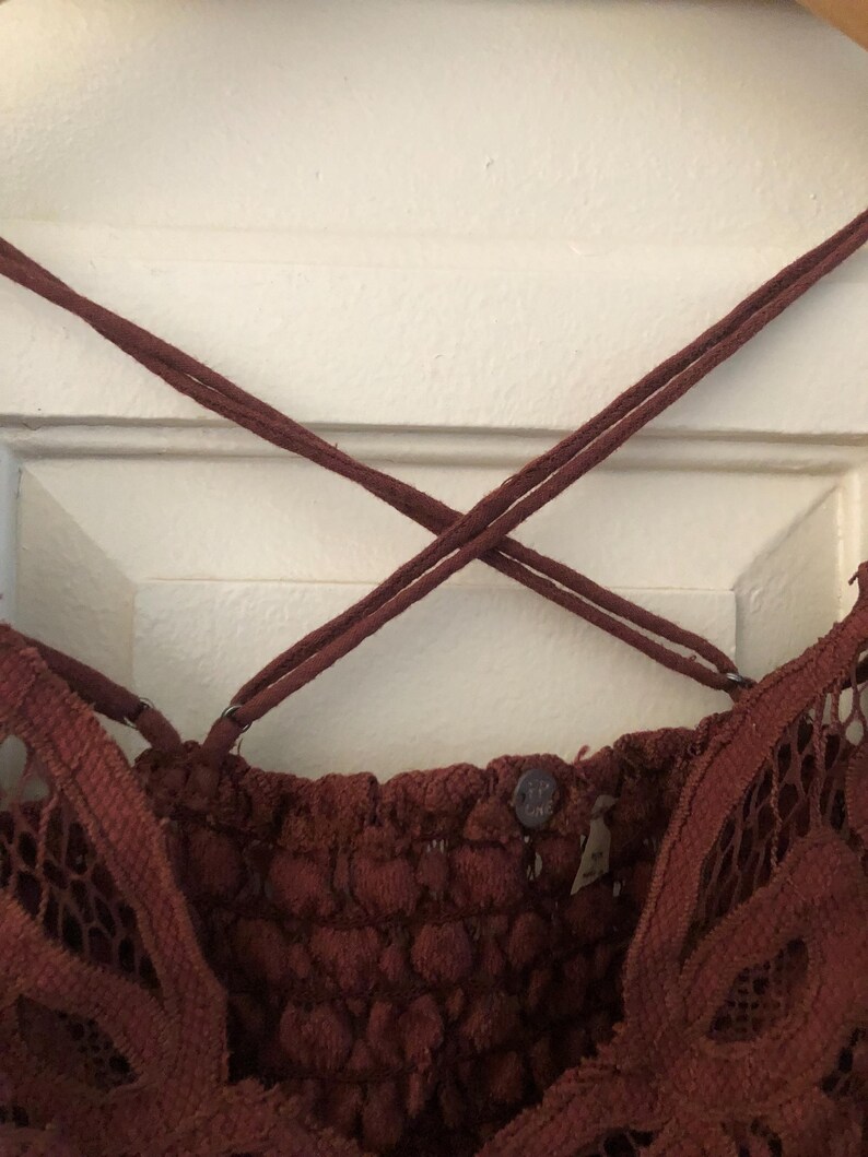 May include: Close-up of a burgundy-colored lace top with crisscross spaghetti straps. The top features a textured, patterned bodice and delicate lace detailing along the edges. A small button and tag are visible.