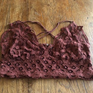 May include: A burgundy-colored lace bralette with a floral pattern. The bralette features a criss-cross back design and a scalloped edge along the bottom. The fabric appears delicate and semi-sheer.