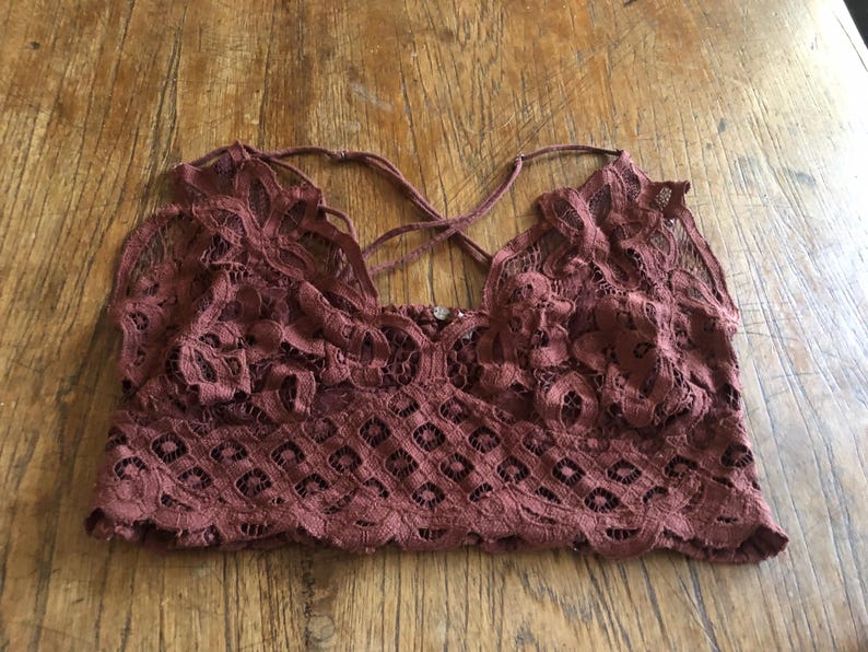 May include: A burgundy-colored lace bralette with a floral pattern. The bralette features a criss-cross back design and a scalloped edge along the bottom. The fabric appears delicate and semi-sheer.
