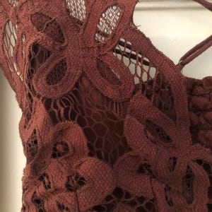 May include: Close-up of a burgundy lace top with floral patterns. The intricate design features a delicate net-like texture and crisscross straps. The top is made of a soft, textured material.