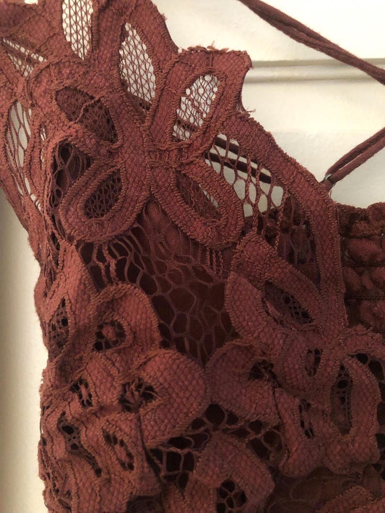 May include: Close-up of a burgundy lace top with floral patterns. The intricate design features a delicate net-like texture and crisscross straps. The top is made of a soft, textured material.