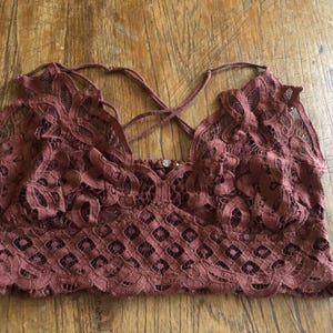 May include: A maroon lace bralette with a criss-cross back design. The bralette features intricate floral and geometric patterns, and thin straps. The garment is made of a soft, delicate lace material.