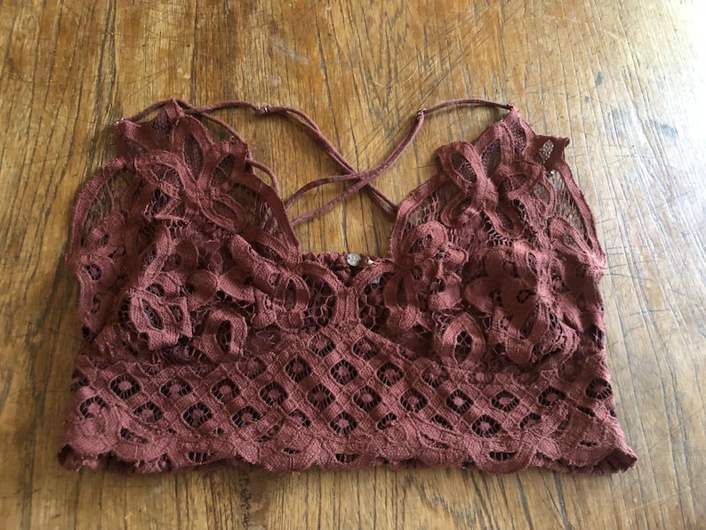 May include: A maroon lace bralette with a criss-cross back design. The bralette features intricate floral and geometric patterns, and thin straps. The garment is made of a soft, delicate lace material.
