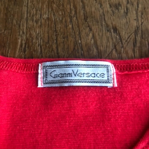 Peut inclure: Gros plan sur un v&ecirc;tement rouge vif avec une &eacute;tiquette Gianni Versace. L'&eacute;tiquette est rectangulaire avec un bord argent&eacute; et le nom de la marque en police serif. Le tissu a un aspect textur&eacute;.