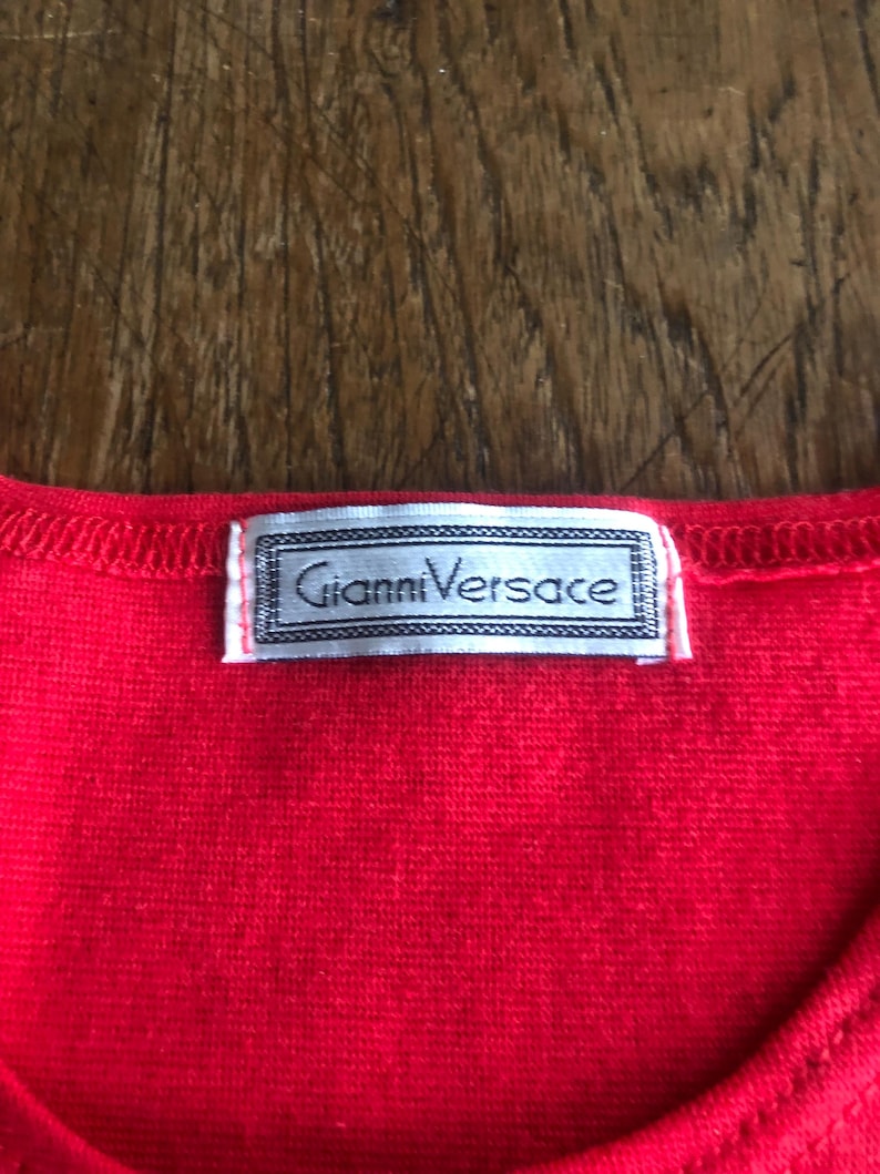 Peut inclure: Gros plan sur un v&ecirc;tement rouge vif avec une &eacute;tiquette Gianni Versace. L'&eacute;tiquette est rectangulaire avec un bord argent&eacute; et le nom de la marque en police serif. Le tissu a un aspect textur&eacute;.