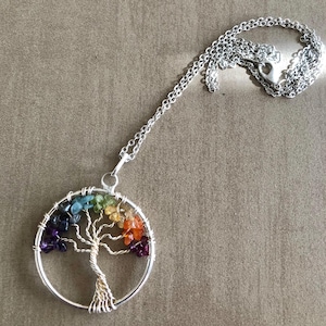 Op de afbeelding: Zilverkleurige ketting met een cirkelvormige hanger met een draadgewikkelde boom. De boom is versierd met gekleurde edelstenen in een regenboogspectrum. De ketting heeft een fijne ketting en een hartvormige sluiting. Diameter hanger: ongeveer 3 cm.