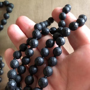 May include: A close-up of several strands of black beaded necklaces. The beads have a faceted, textured surface and are strung on black cord. The necklaces are displayed against a neutral background.