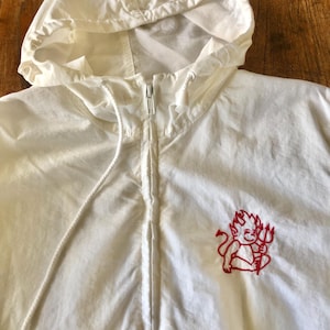May include: A white zip-up hooded jacket featuring a red embroidered graphic of a small devil character holding a trident on the left chest. The jacket includes a full-length front zipper, a drawstring hood with white cords, and long sleeves.